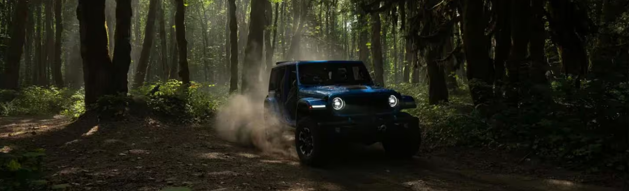 A blue Jeep Wrangler 4xe drives through a dusty forest trail.